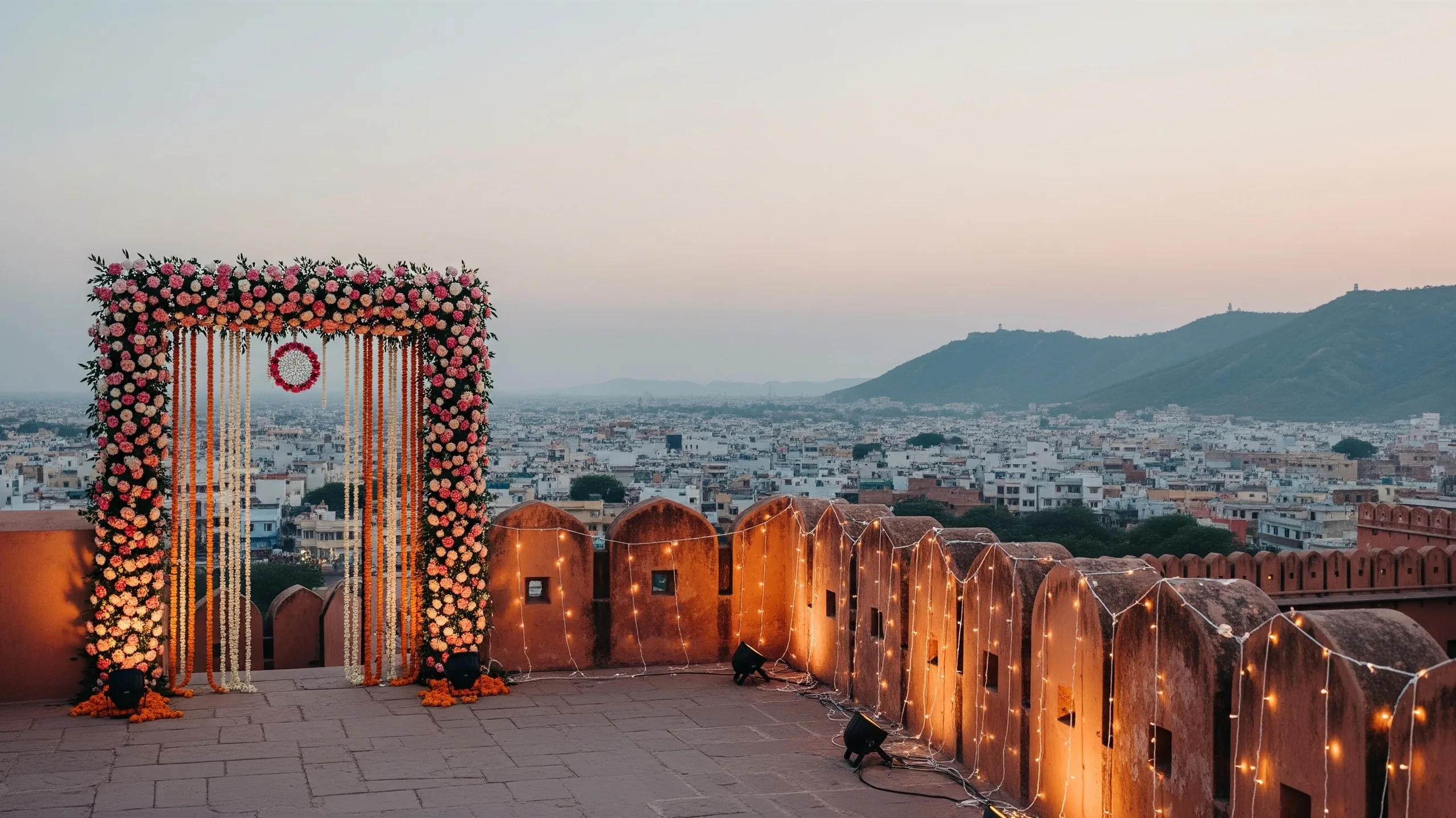 An atmospheric wide view of a Jaipur fort rampart at dusk with a decorated floral gateway and fairy lights, the city and hills behind, serene true-to-life light, no signage, styled view